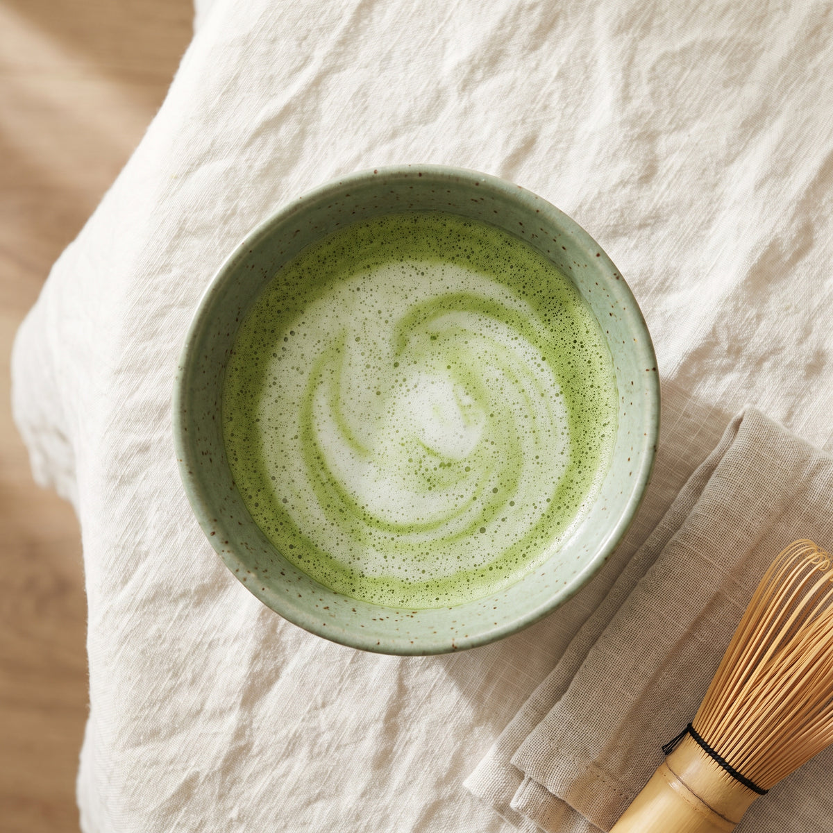 Whisked Mind Full Matcha latte in a sage-green speckled ceramic bowl on cream linen, seen from above with a bamboo whisk alongside