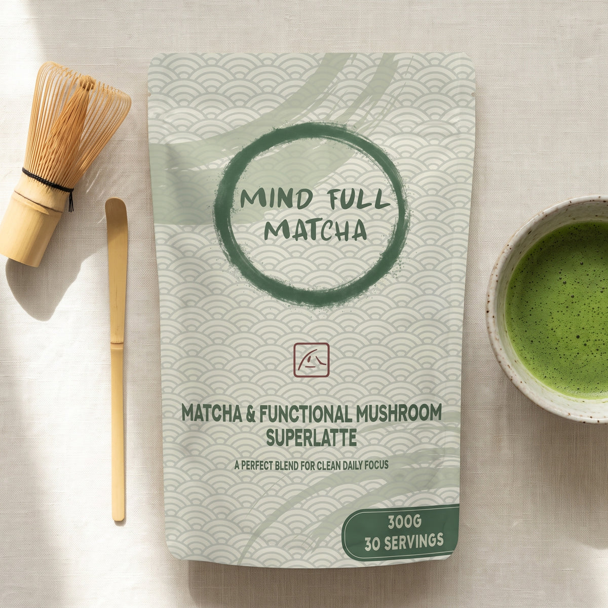 Flat lay of the Mind Full Matcha 300g pouch with a bamboo chasen whisk, a bamboo chashaku scoop and a speckled ceramic bowl of whisked matcha on cream linen